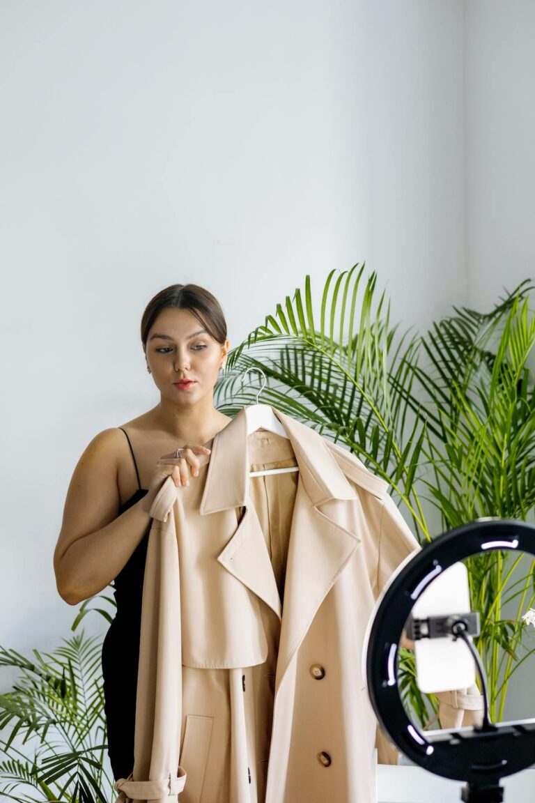 A fashion vlogger presents a chic beige coat during an indoor recording session with ring light.
